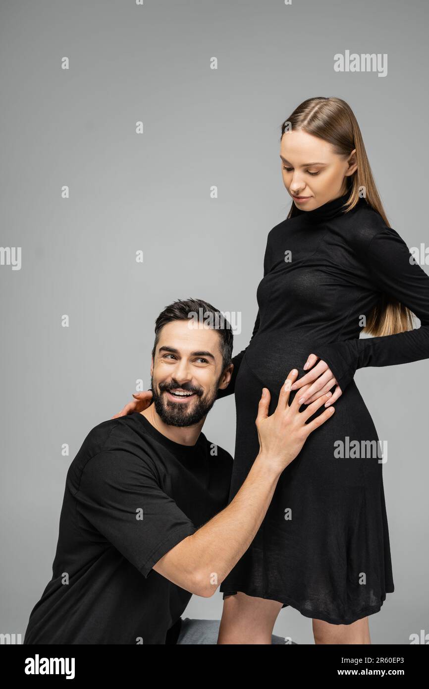 Smiling bearded man in t-shirt listening belly of trendy and pregnant wife with natural makeup in stylish black dress while standing isolated on grey, Stock Photo