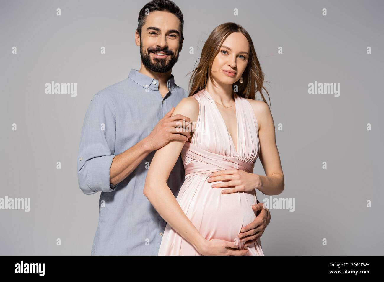 Portrait of positive and bearded man hugging elegant pregnant wife in ...