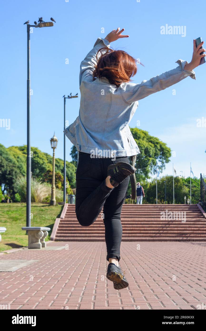 back view of young woman jumping in the park, dressed in a blue jean jacket and black jean pants. Stock Photo