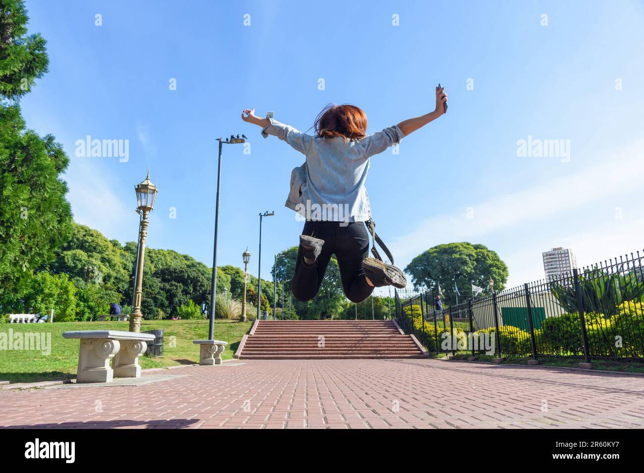 rear view of young woman jumping in the park with her legs and arms open, she is dressed in a jean jacket and pants, red hair, and the sky in the back Stock Photo