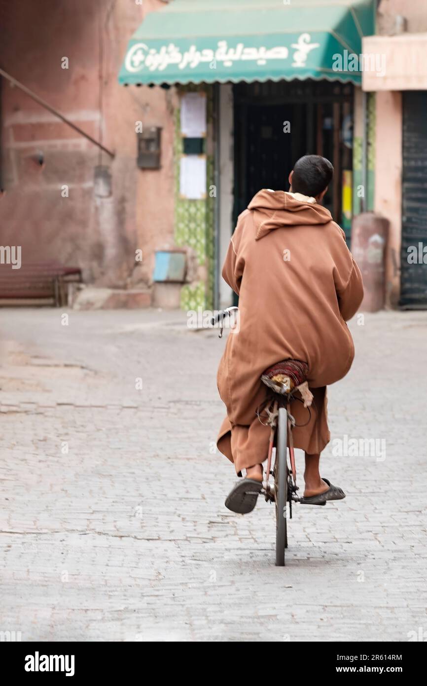 A male cycling along a cobbled alleyway in the Medina Marrakesh, Morocco. The man is wearing a Berber Caftan Brown Djellaba with a baggy hood or qob Stock Photo