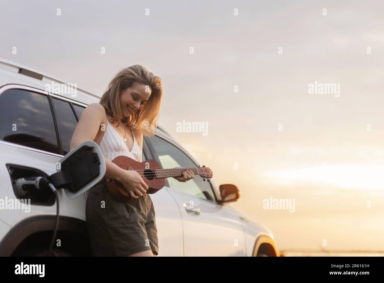 Portrait of young woman leaning on her car,playing on ukulele and enjoying summer time. Stock Photo