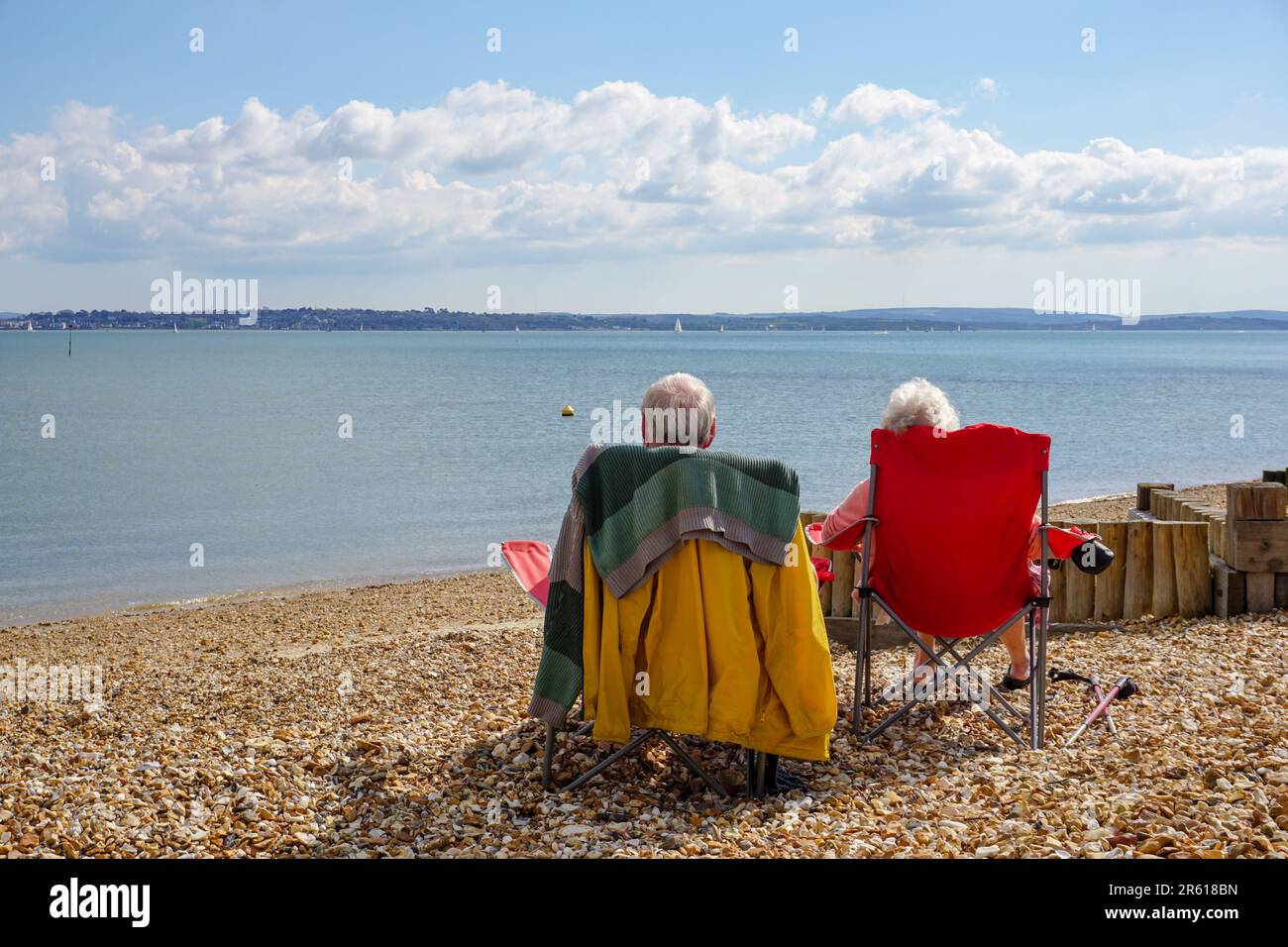 Elderly couple relaxing on stony beach. A married couple sitting on deckchairs looking out to sea on sunny day. Togetherness concept Stock Photo