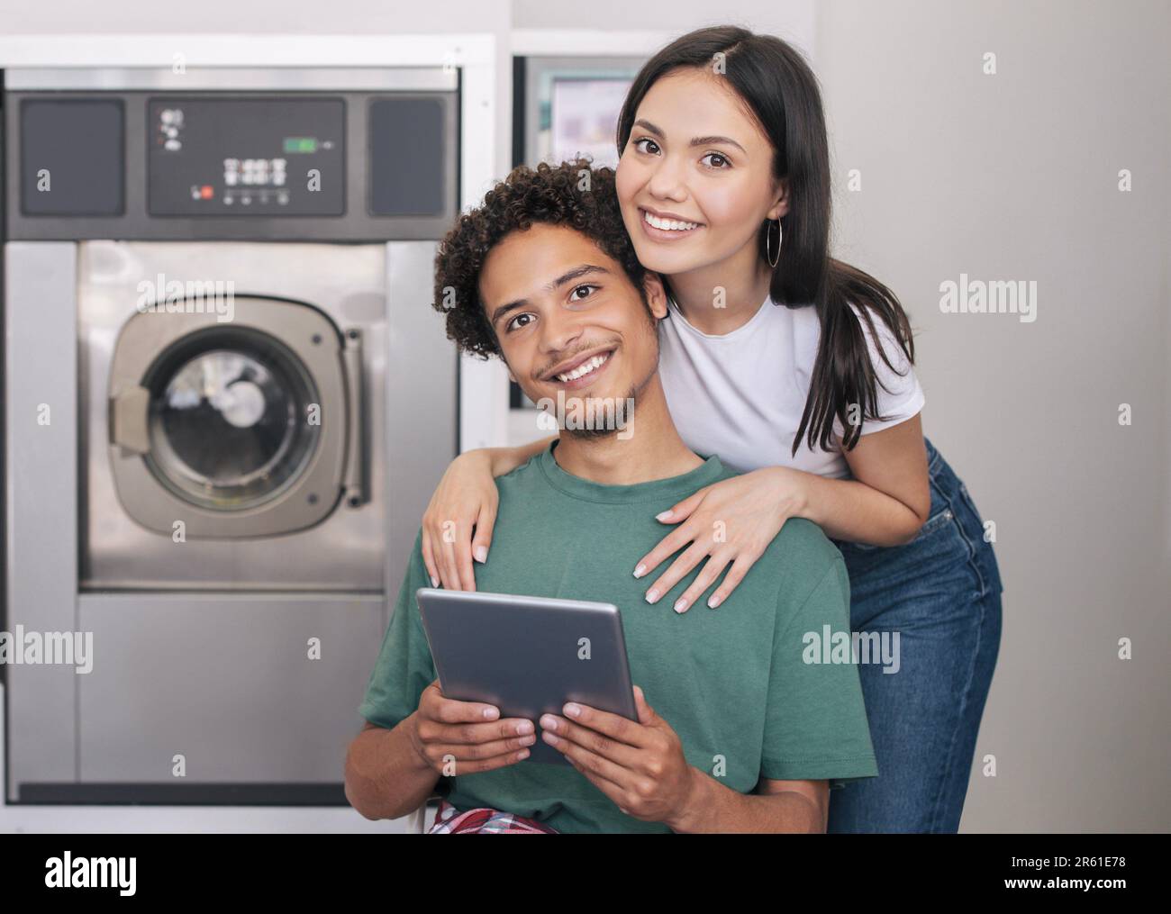 Diverse Couple Posing With Tablet Near Washing Machine At Laundry Stock Photo