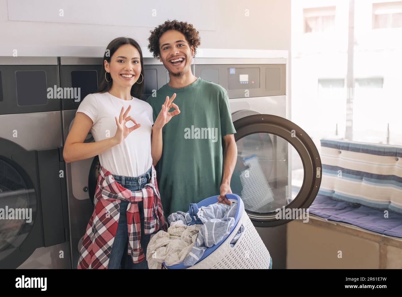 Couple Gesturing OK Posing Near Laundromat Washing Machines At Laundry Stock Photo