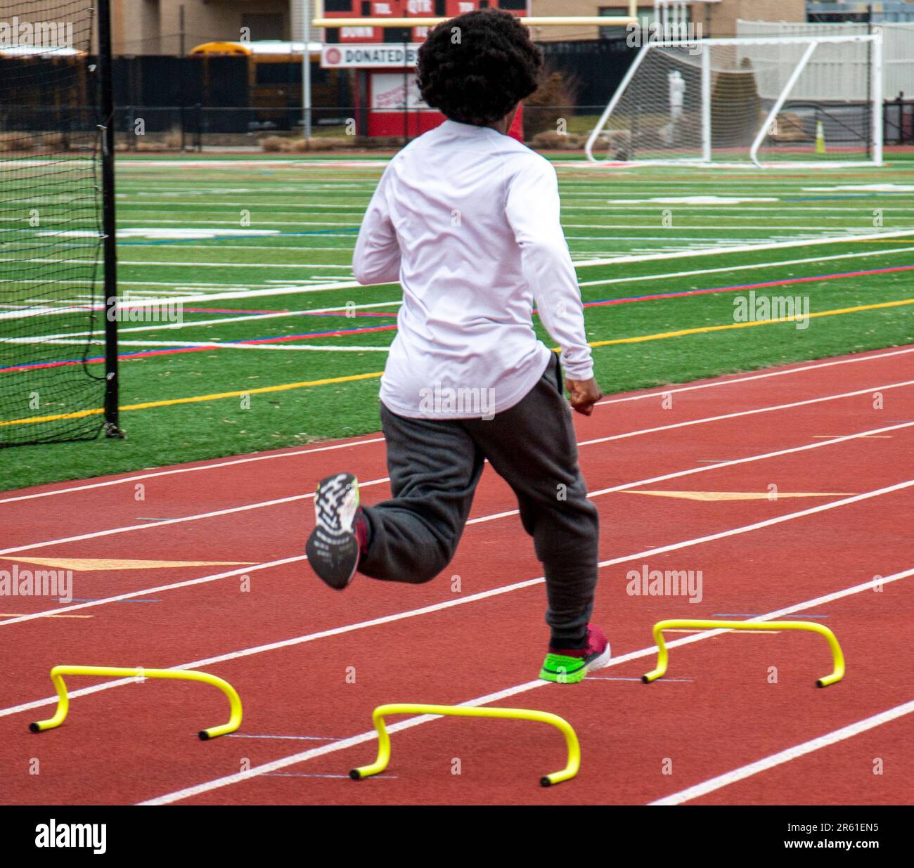 Rear view of an African American track runner sprinting over six inch hurdles on a red track in lane during speed and agility practice. Stock Photo