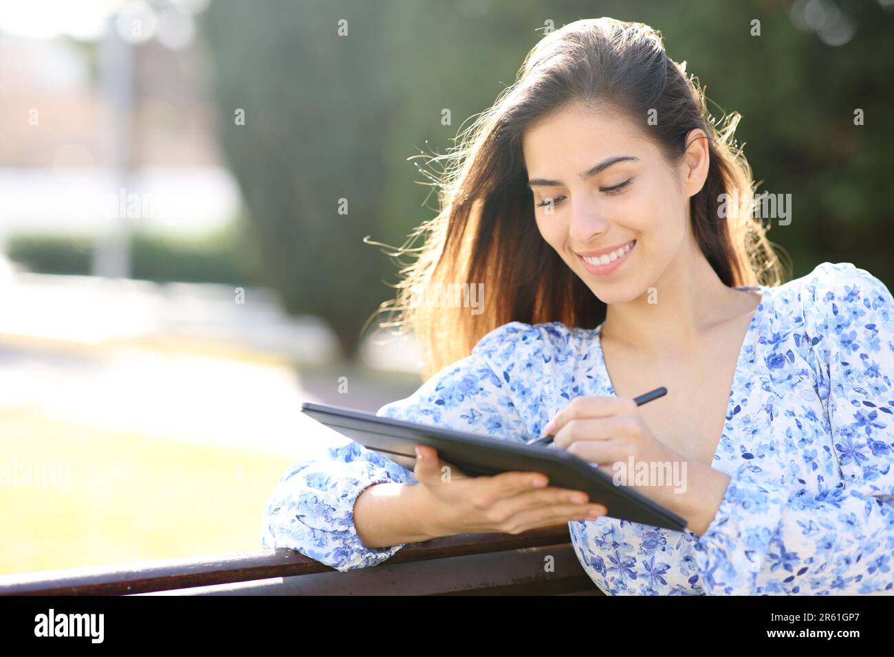 Happy woman writing on tablet with pen sitting in a park Stock Photo