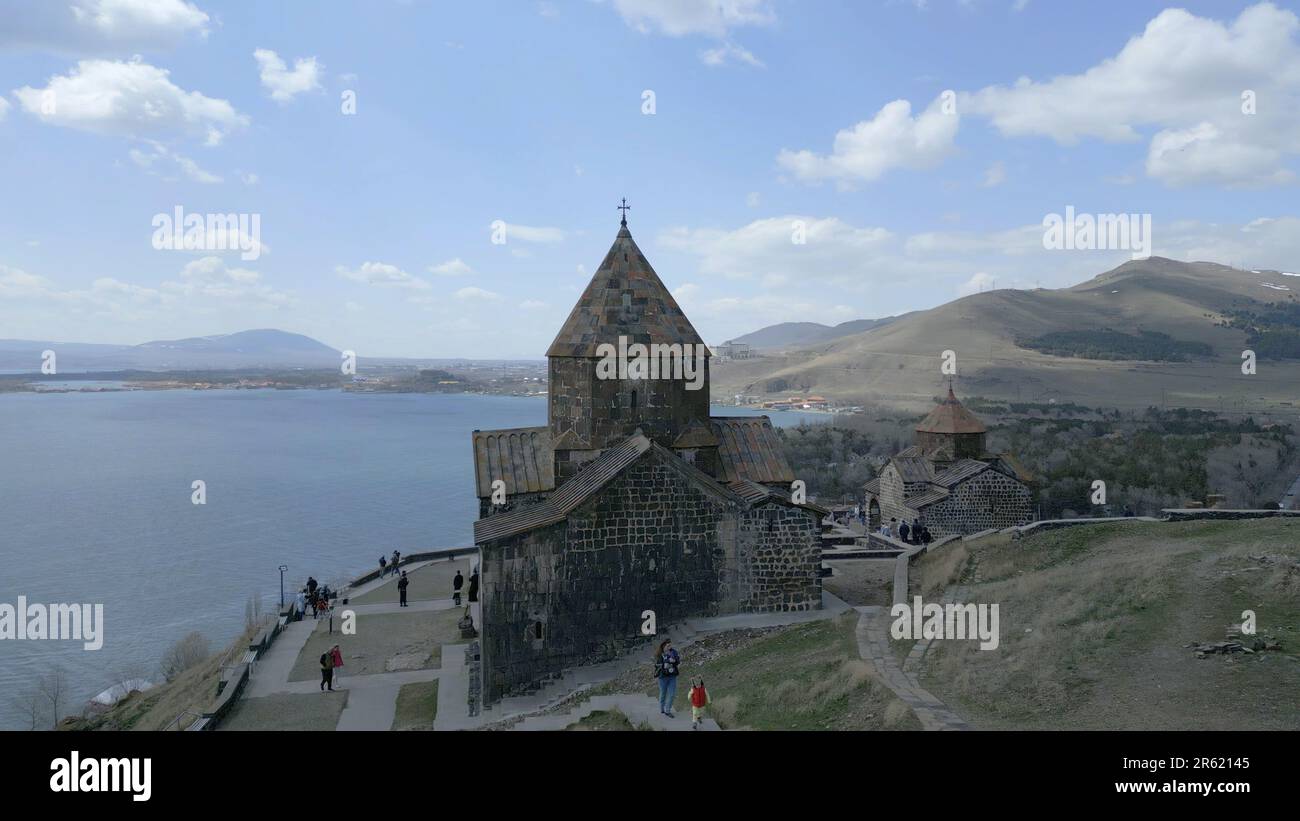 The Sevanank Monastery Complex in Armenia. Monastery buildings with crosses, on the background of Lake Sevan, houses, people walking, high snowy mount Stock Photo