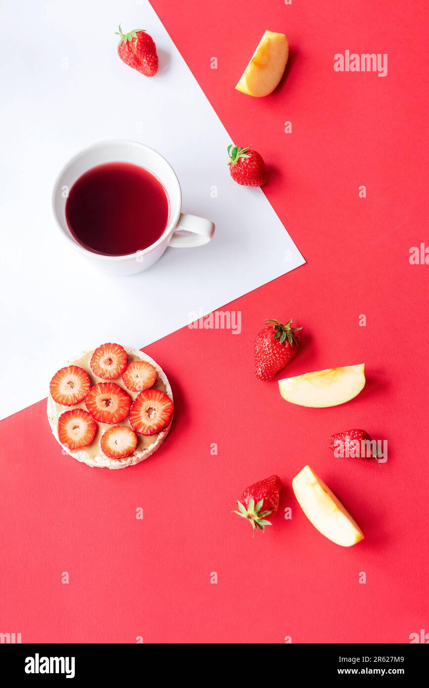 Cup of fruit tea, strawberry sandwich, apple slices on red and white background. Top view. Stock Photo