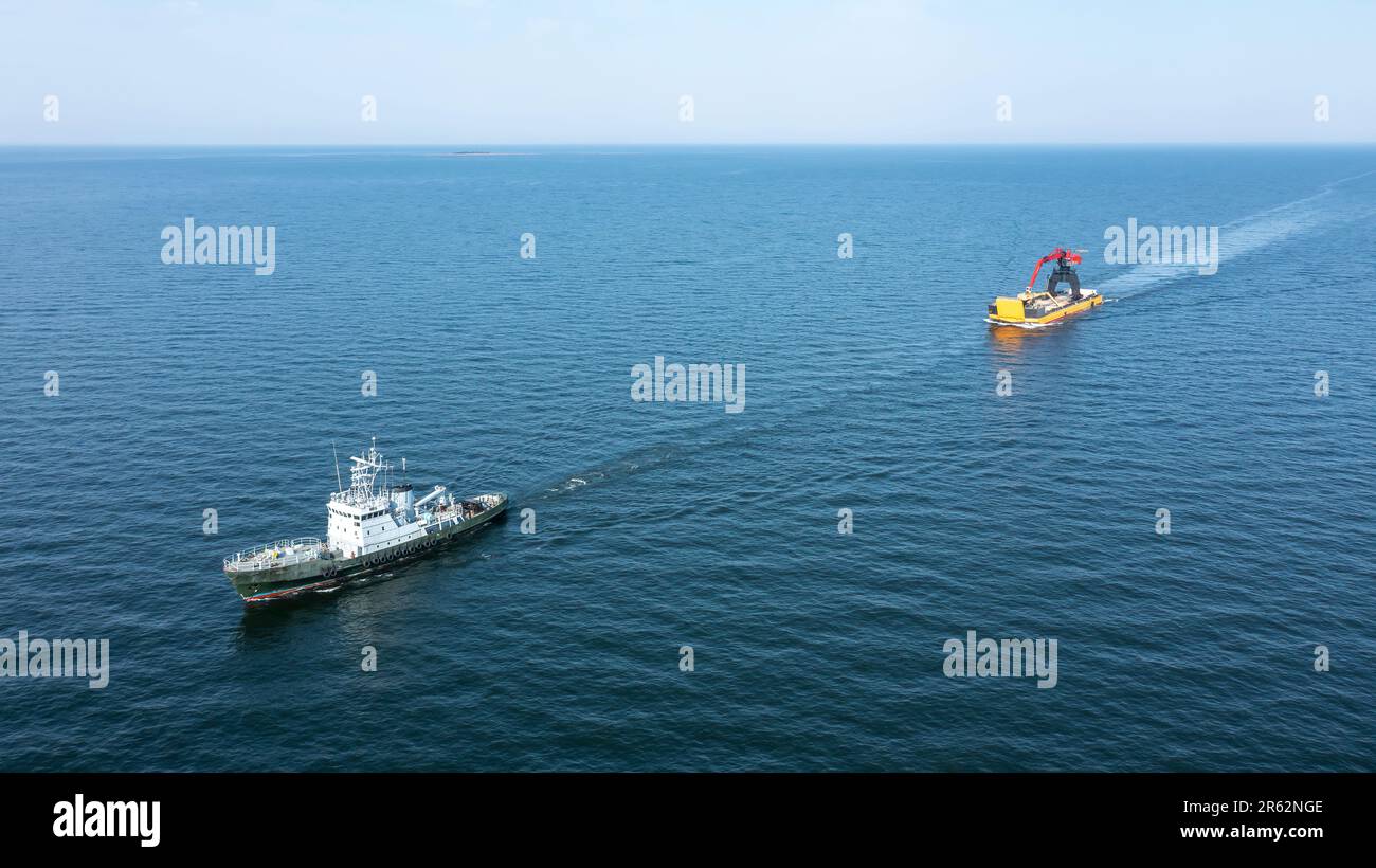 Tug boat travelling across the open ocean pulling a yellow barge loaded with tracked harbour material handling cranes. Aerial front view. Stock Photo