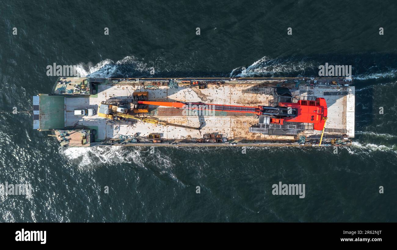 Barge loaded with tracked harbour material handling cranes, being pulled across the open ocean. Aerial top down view. Stock Photo