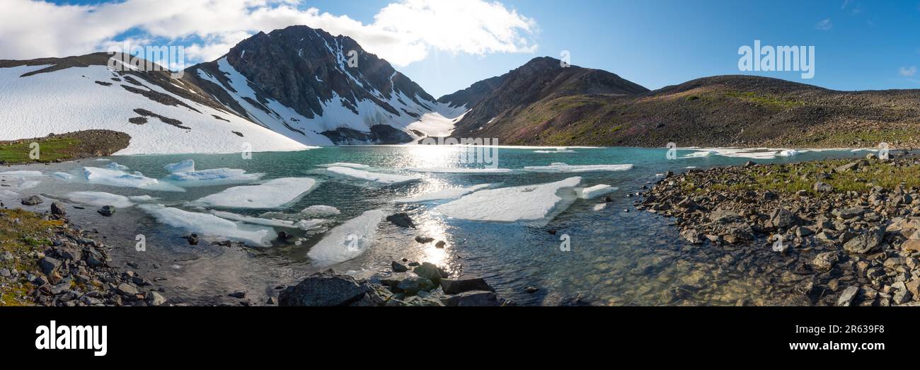 Back country camping area in northern arctic Canada during summer time with panoramic view over glacial lake, yellow tent, huge mountains, snow, ice Stock Photo