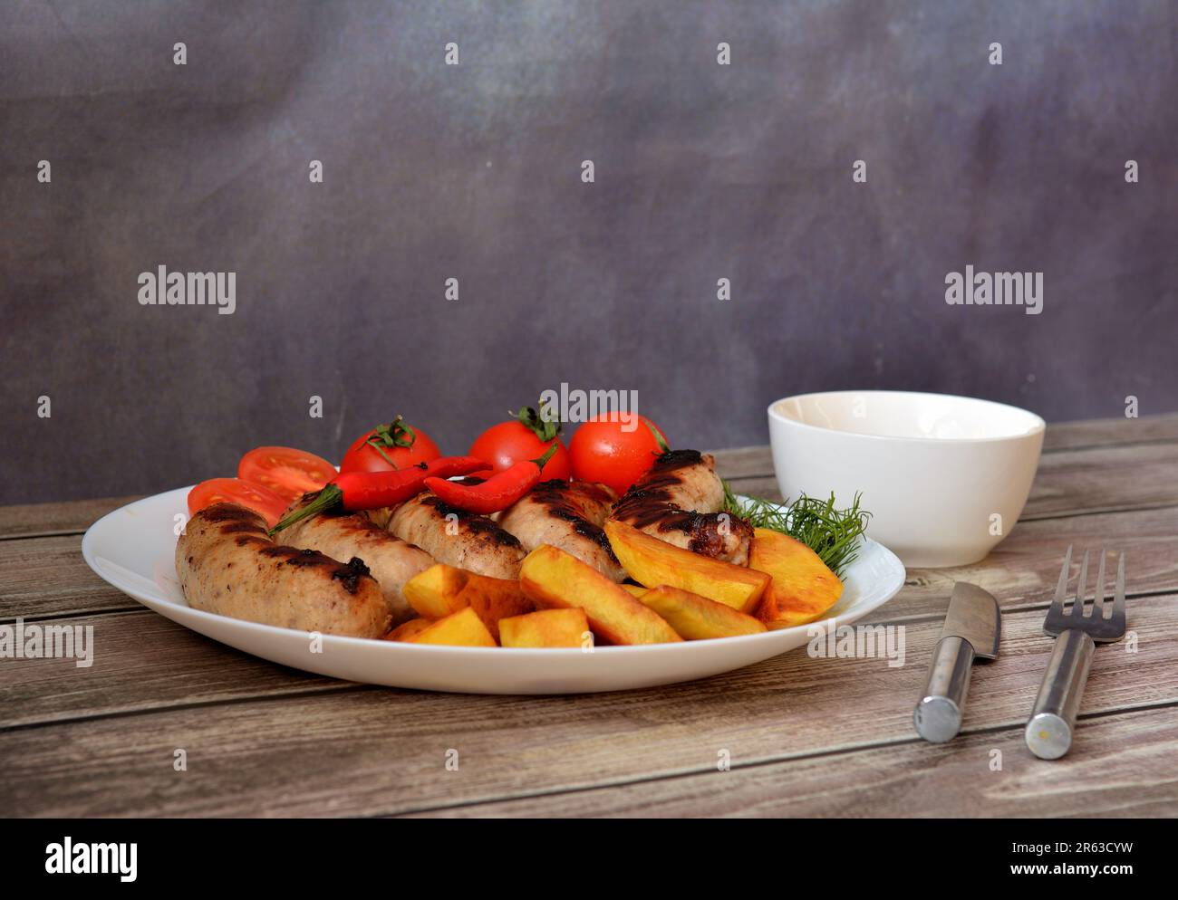 A white ceramic plate with grilled sausages, cherry tomatoes, fried potatoes and herbs on a wooden table, next to it is a gravy boat, a fork and a kni Stock Photo