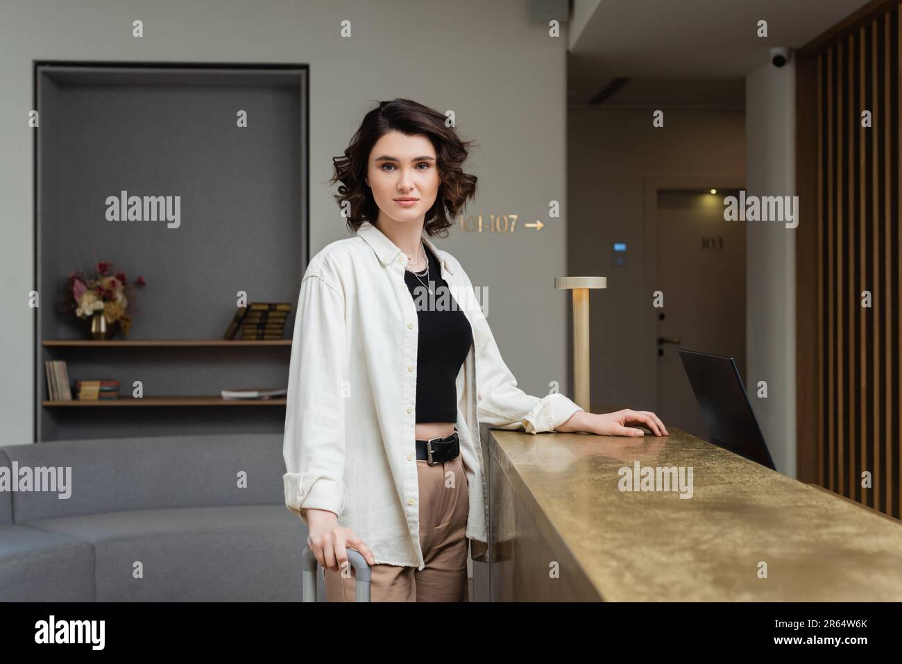 confident woman in black crop top, white shirt and beige pants, with wavy brunette hair standing near lamp on reception desk and looking at camera in Stock Photo