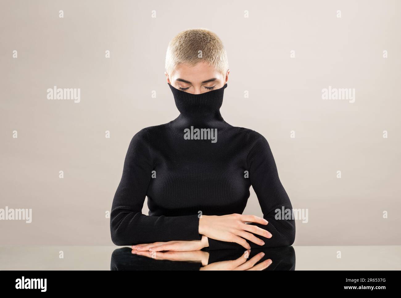 Calm young female model with short blond hair covering mouth with collar of black turtleneck while sitting at mirrored table with closed eyes against Stock Photo