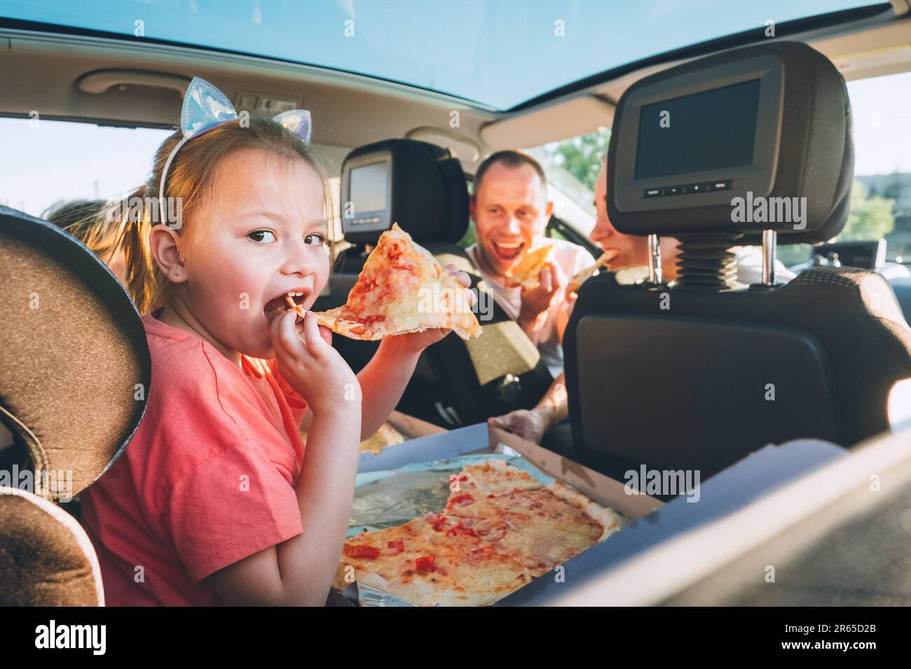 Little girl with open mouth portrait eating Italian pizza sitting in modern car with mother and father. Happy family moments, childhood, fast food eat Stock Photo