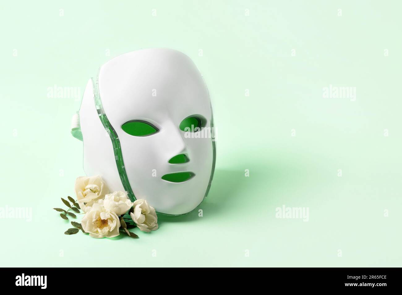 Cosmetic LED facial mask regenerative treatment on a green background with delicate rose flowers and a place for your text. Health and beauty of the f Stock Photo
