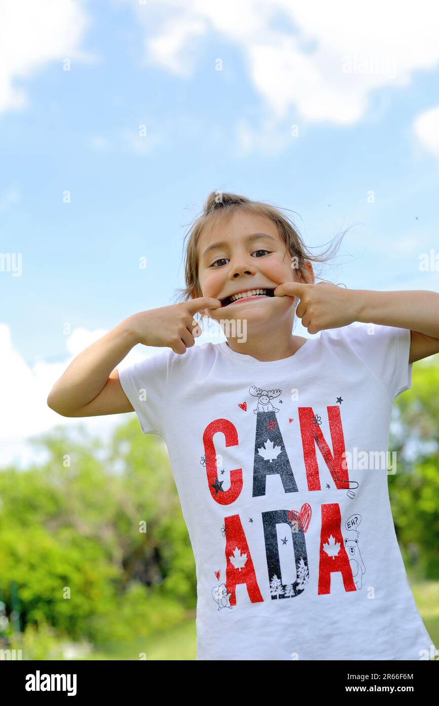 6 year old girl making a funny face while enjoying canada day. canada shirt. blue sky background. Stock Photo