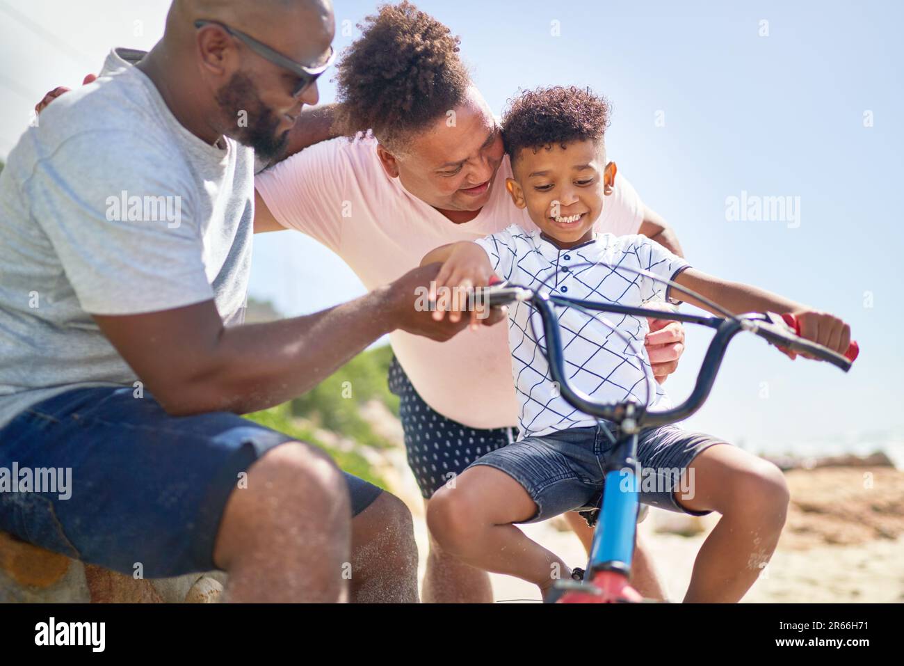 Happy gay male couple helping son riding bike on sunny beach Stock Photo