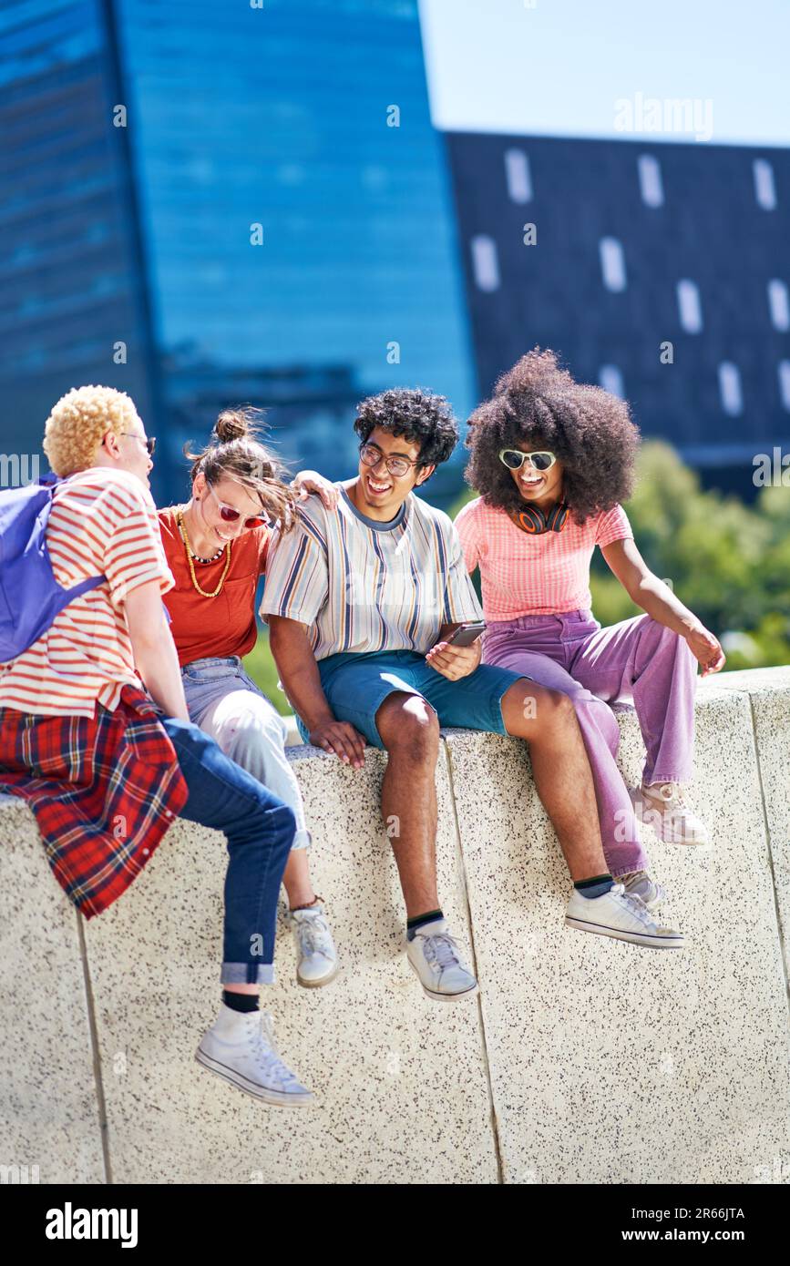 Young friends laughing, hanging out on ledge in sunny city Stock Photo