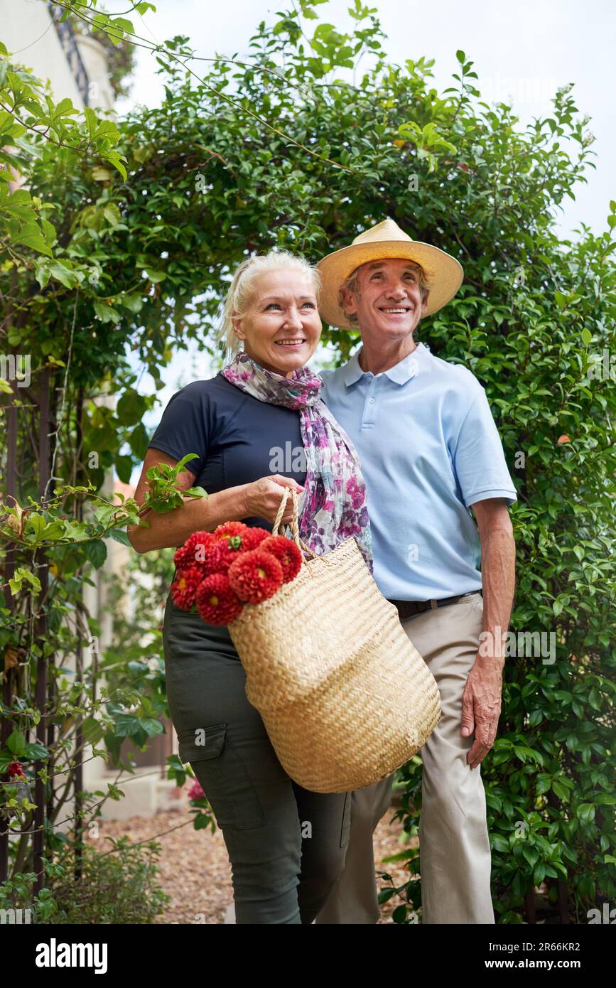 Happy senior couple with flowers walking under trellis in garden Stock Photo