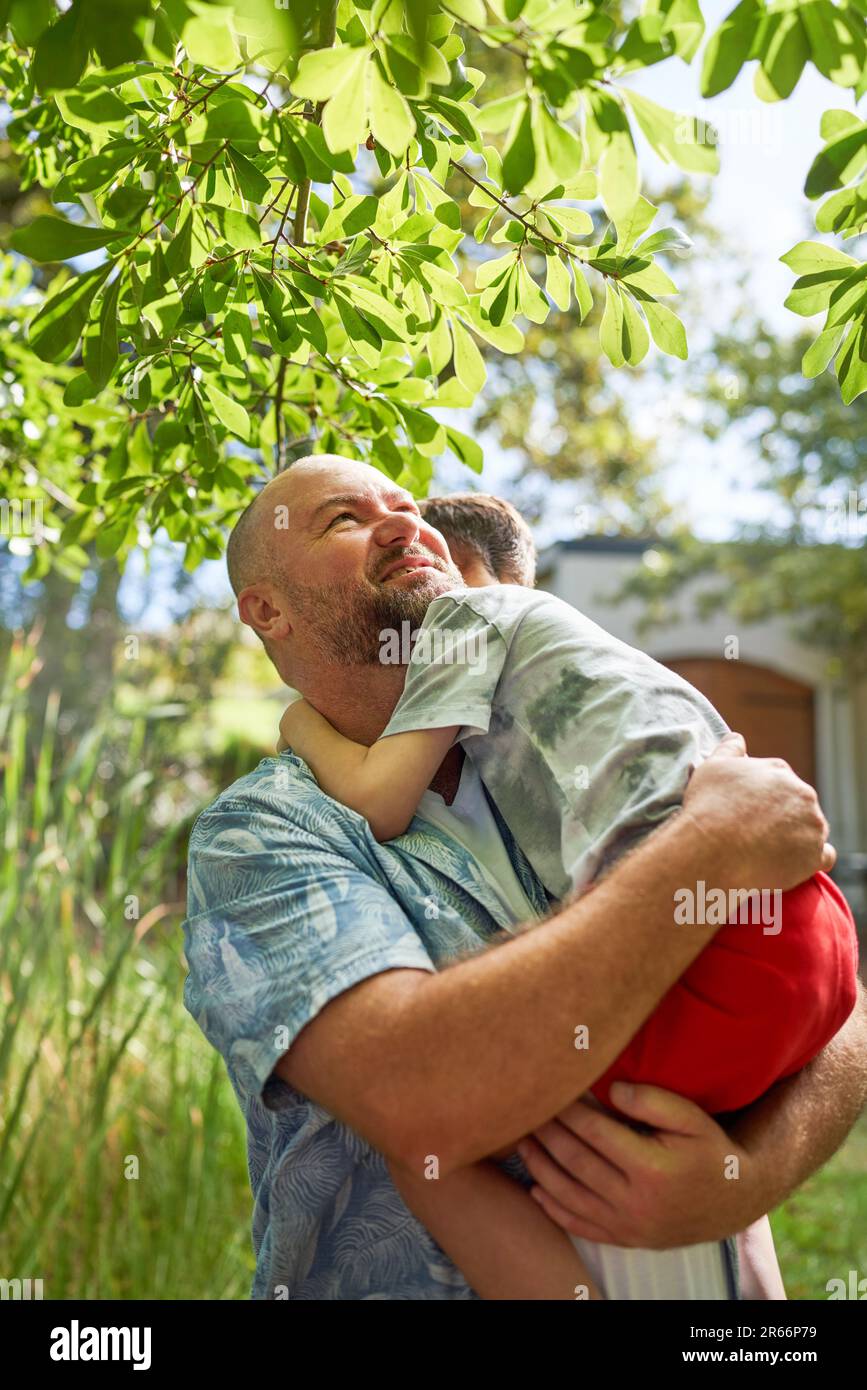 Happy father holding, hugging son under tree in summer backyard Stock Photo