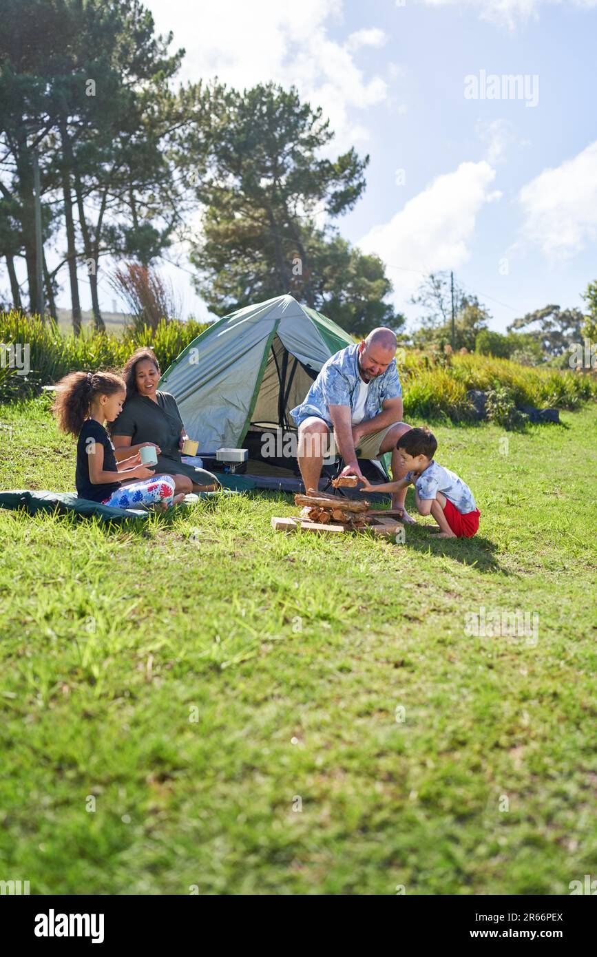 Happy family camping, stacking firewood outside tent in sunny grass Stock Photo