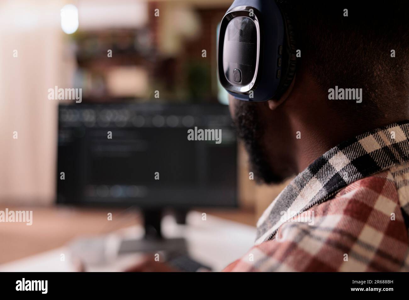 Closeup on freelance coder with wireless headphones listening to music while developing software on personal computer at home. African american programmer writing code while working remote on pc. Stock Photo