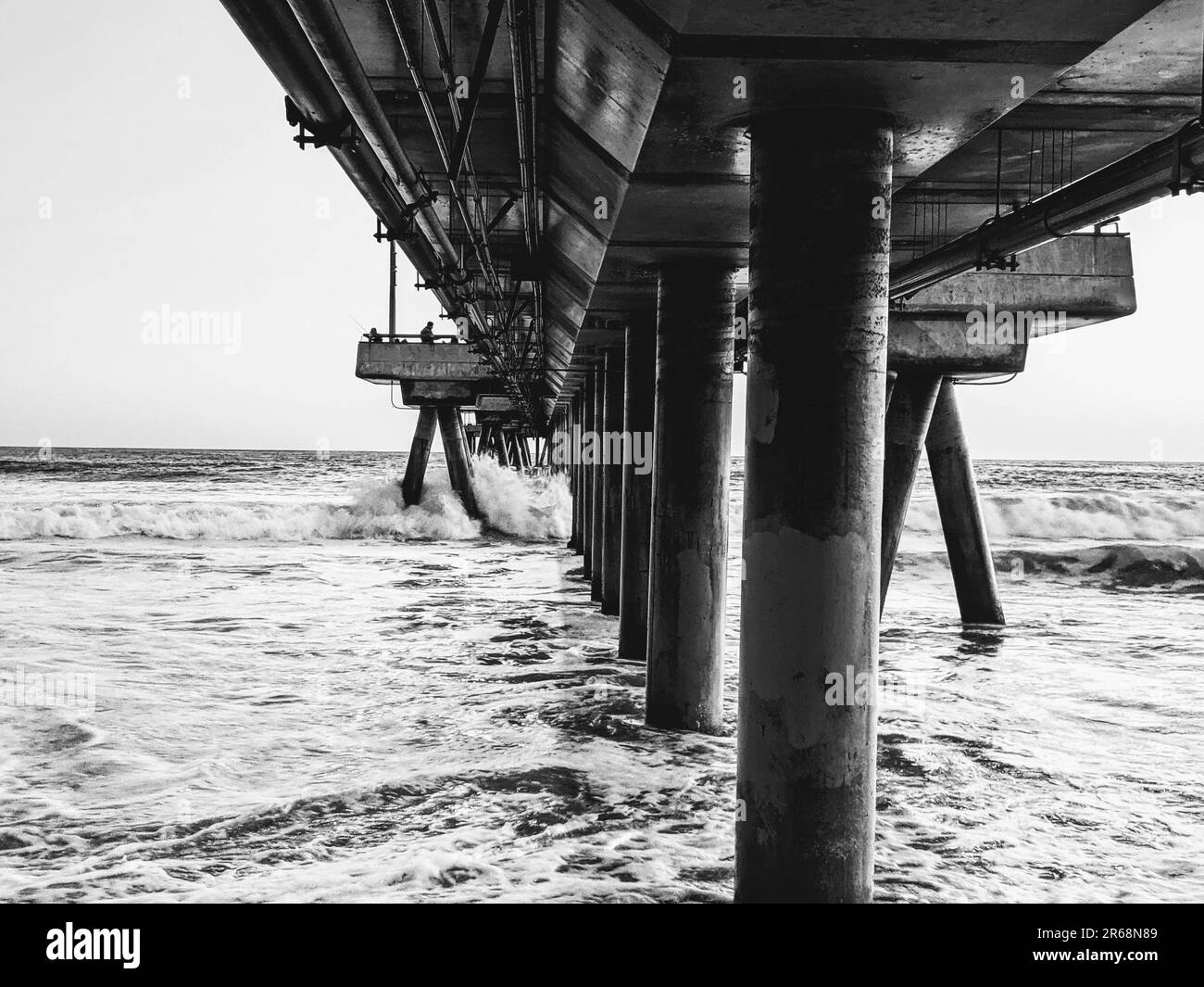 A grayscale shot of waves crashing into a pier on Venice Beach Stock Photo