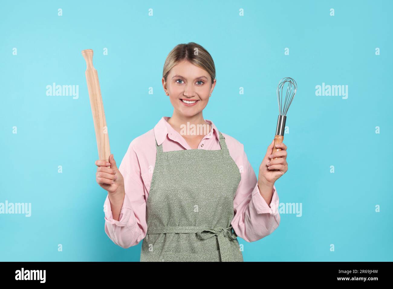 Beautiful young woman in clean apron with kitchen tools on light blue ...