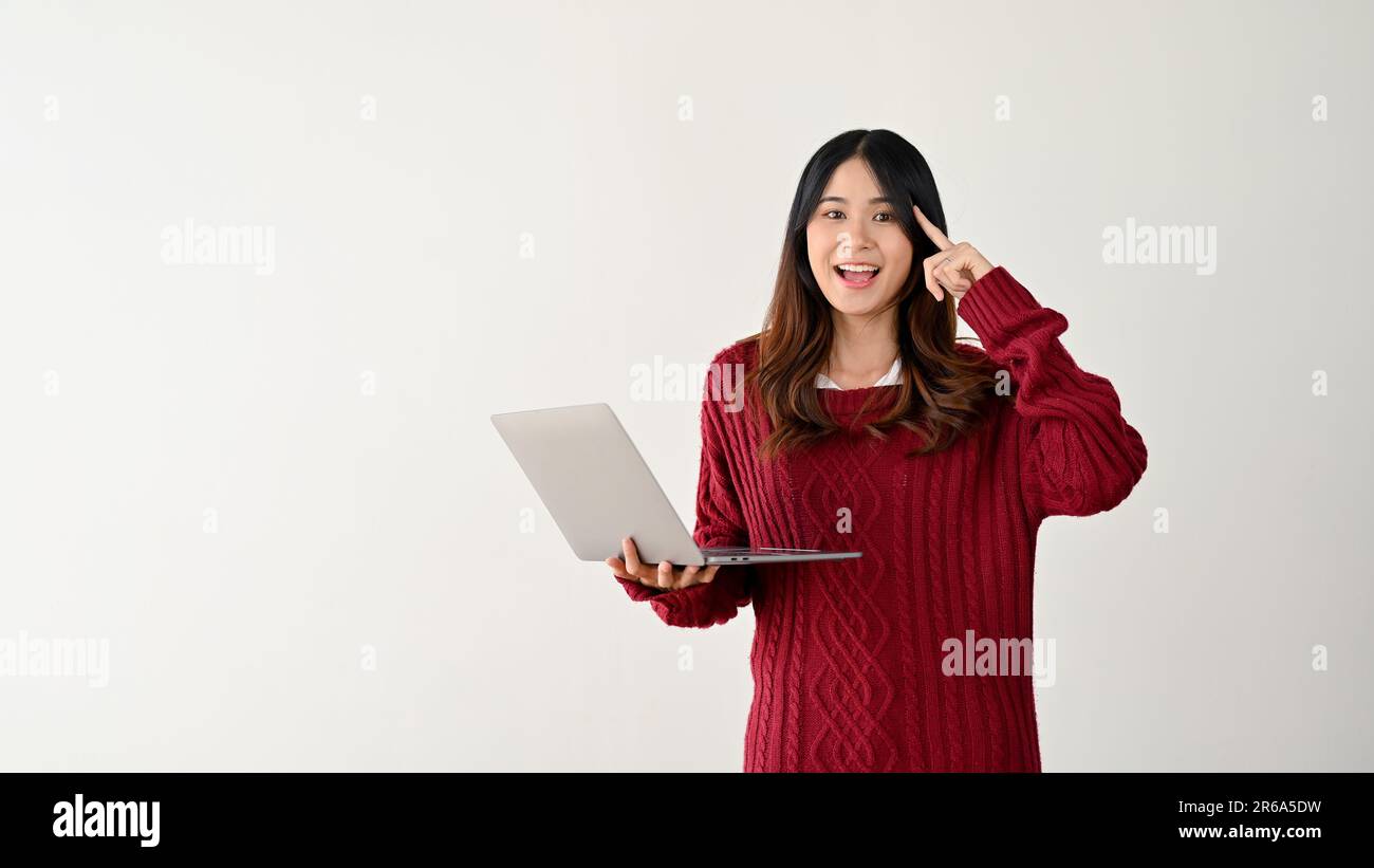 A positive and smart young Asian female college student is pointing her finger at her head while holding her laptop and standing against an isolated w Stock Photo