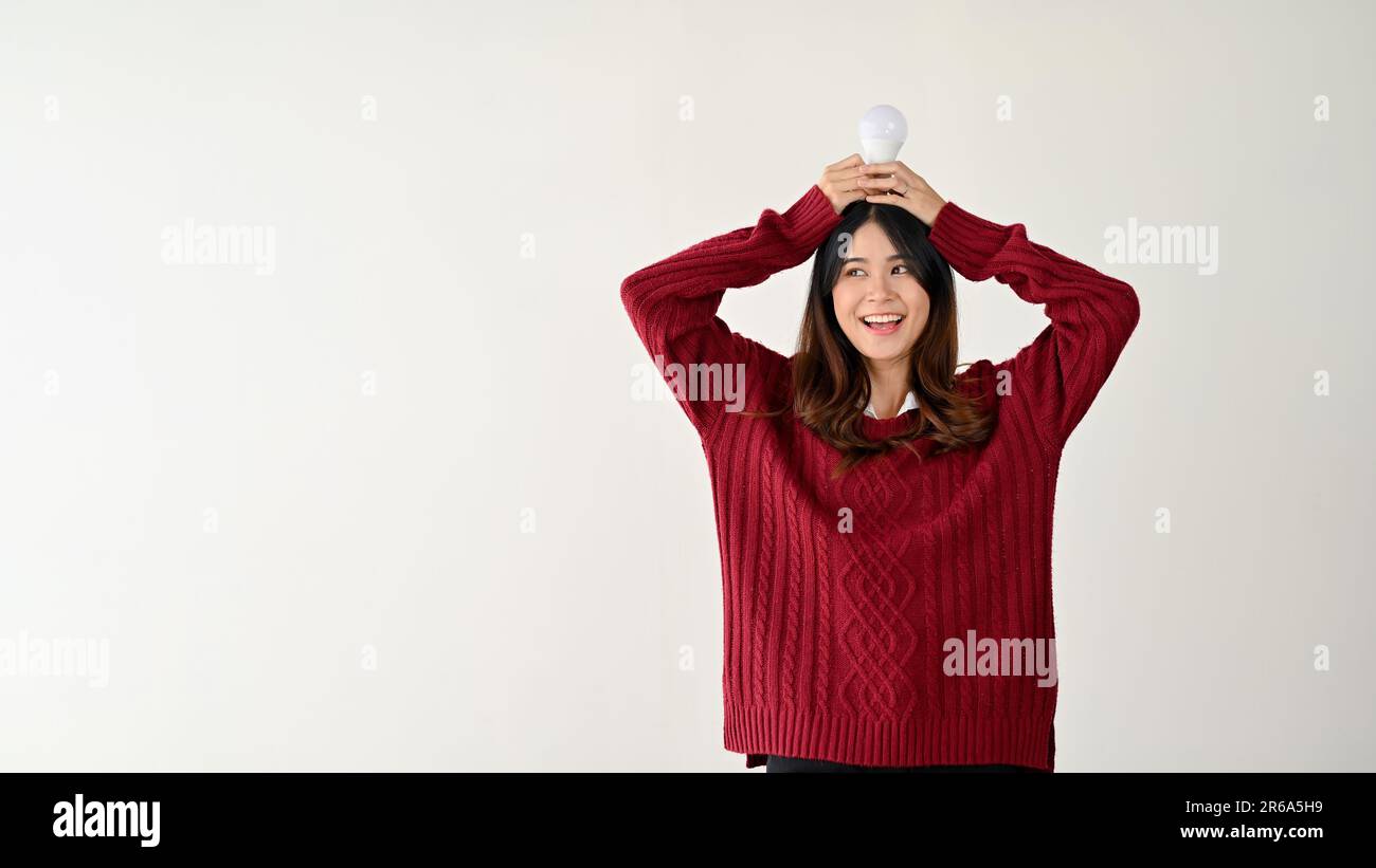 A cheerful and positive young Asian female is putting a light bulb on her head and looking aside at a copy space on a white background. idea, creative Stock Photo