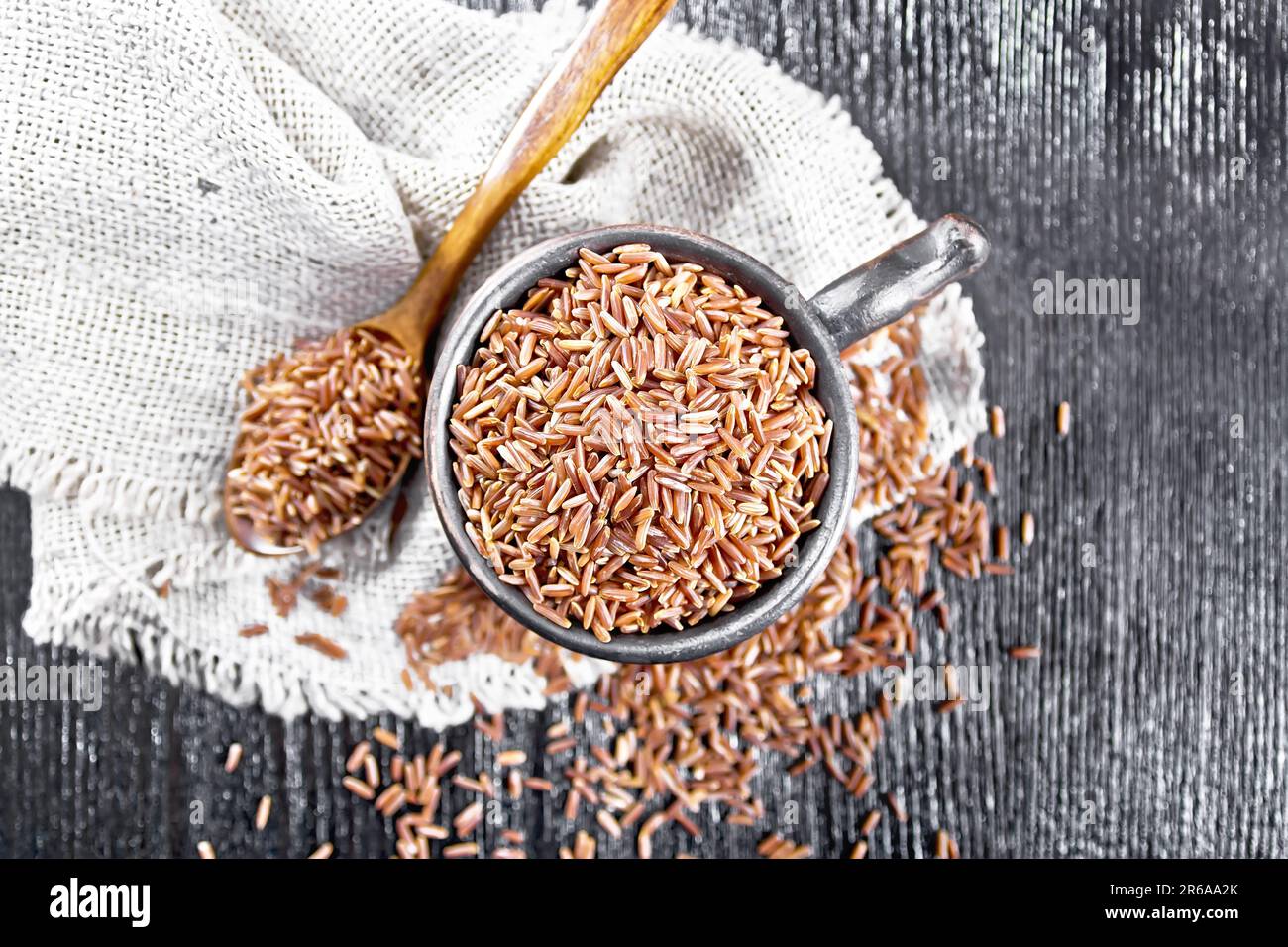 Red raw rice in clay cup and a spoon on a burlap napkin on background of wooden board from above Stock Photo
