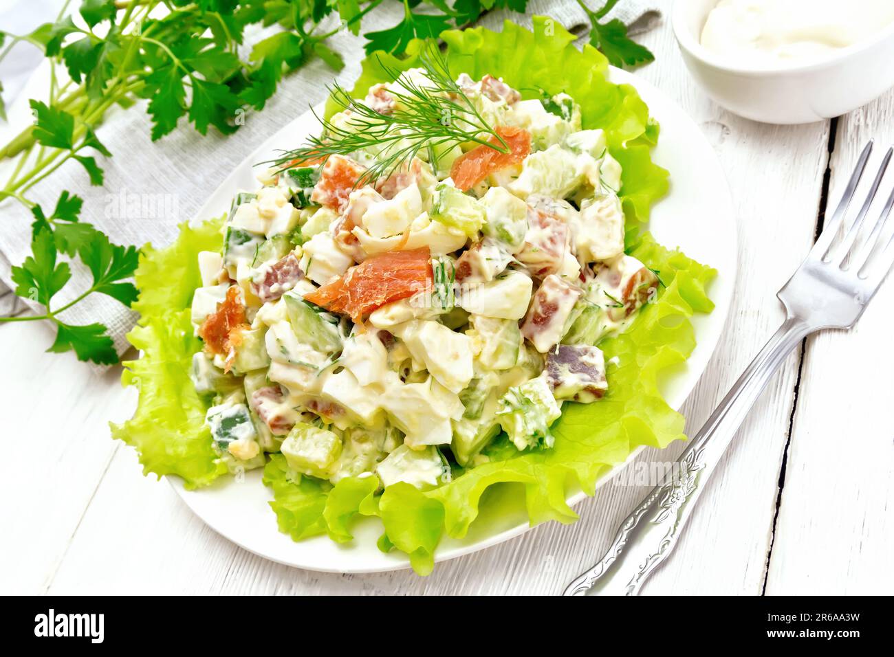 Salad from salmon, cucumber, eggs and avocado, dressed with mayonnaise on lettuce leaves in a plate, kitchen towel, dill, parsley and fork on a wooden Stock Photo