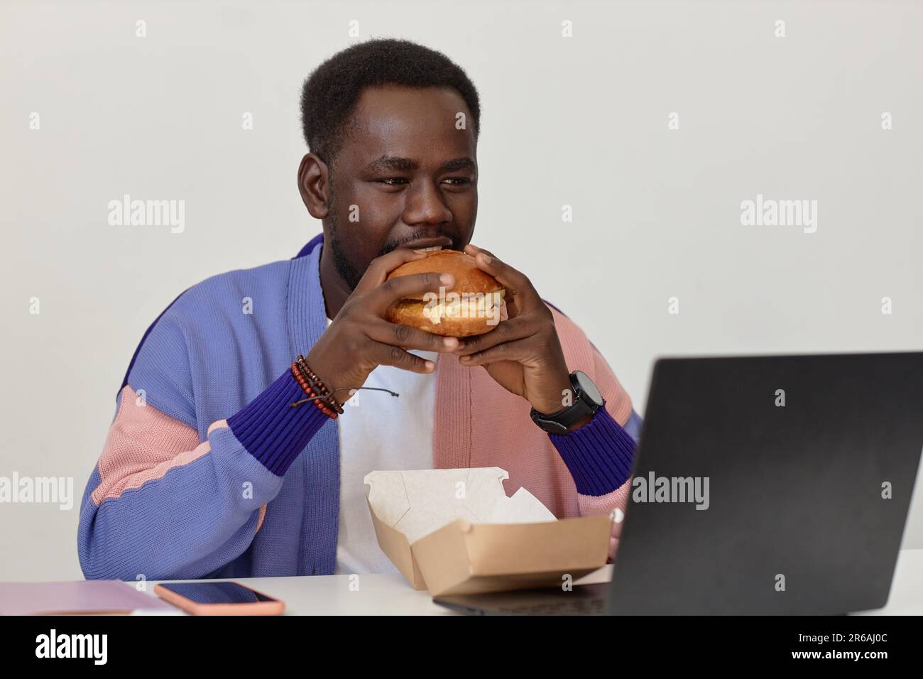 Portrait of young black man eating takeout burger at workplace in office against white wall, copy space Stock Photo
