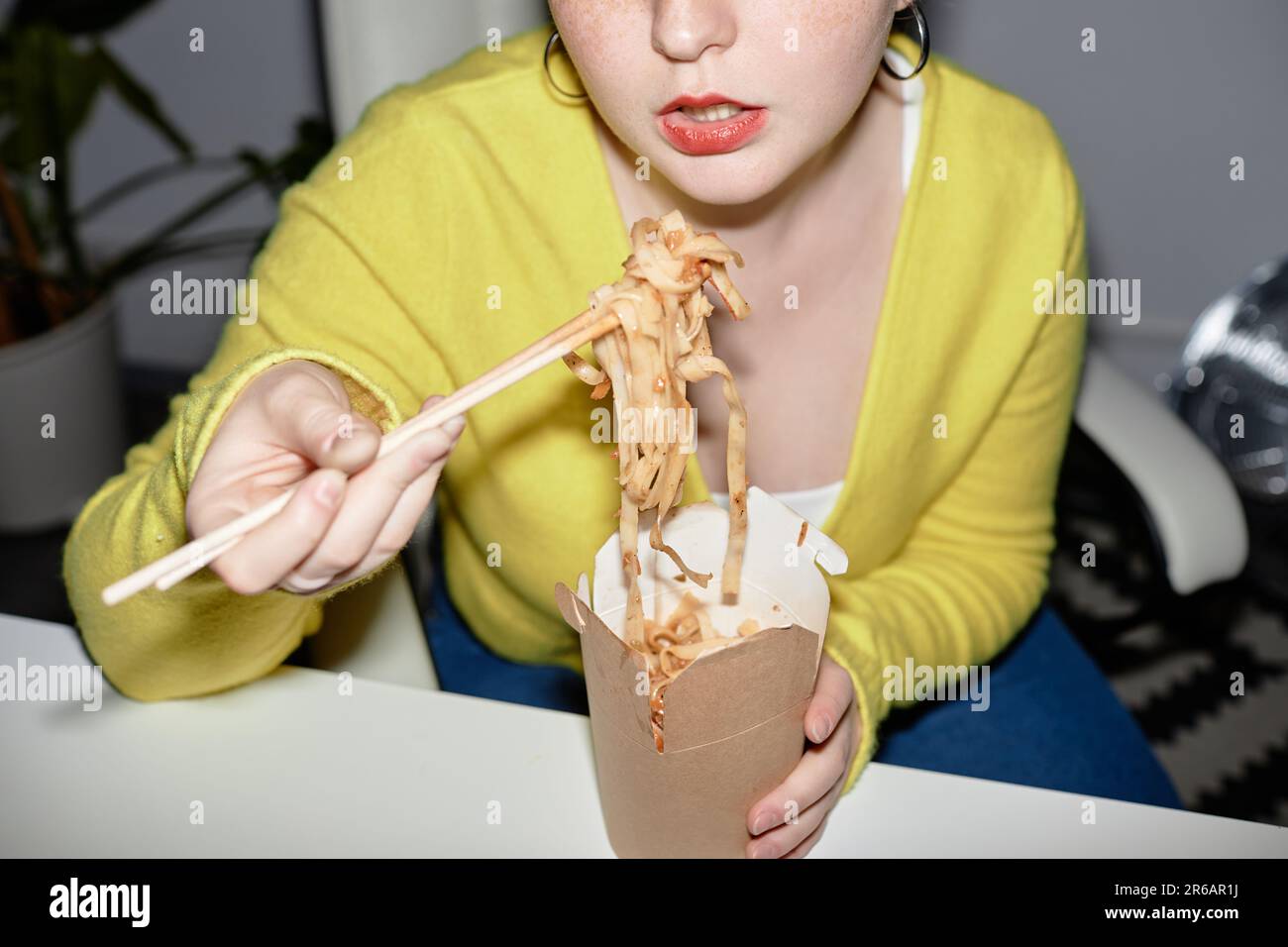 Closeup of young woman eating takeout noodles indoors, shot with flash Stock Photo