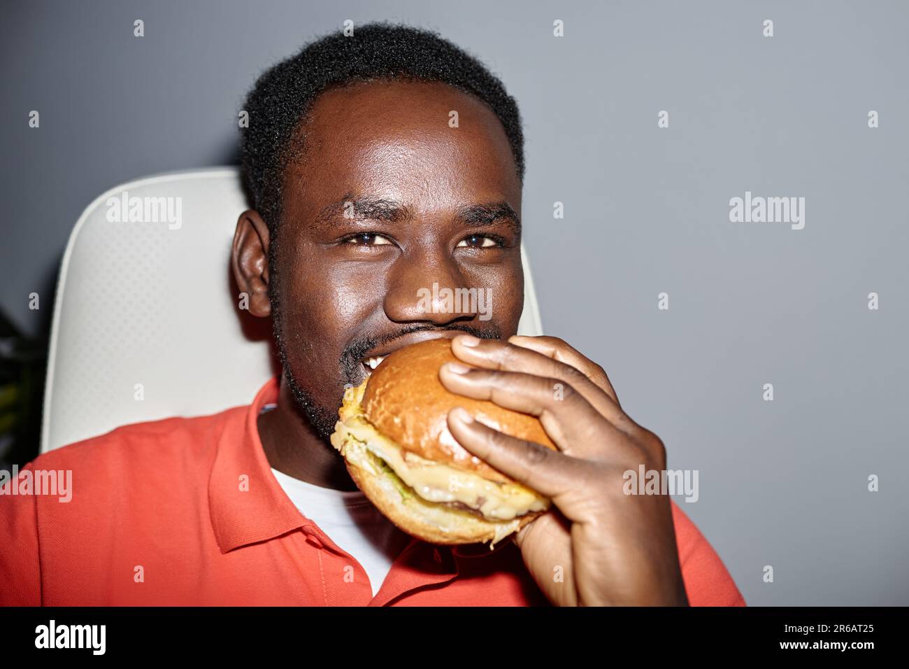 Closeup portrait of black man eating burger indoors and looking at camera happily shot with flash, copy space Stock Photo
