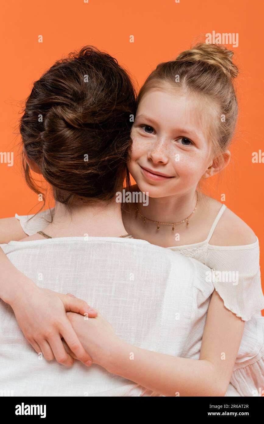 family moments, happy preteen girl hugging brunette mother on orange background, looking at camera, white sun dresses, female bonding, love and care, Stock Photo