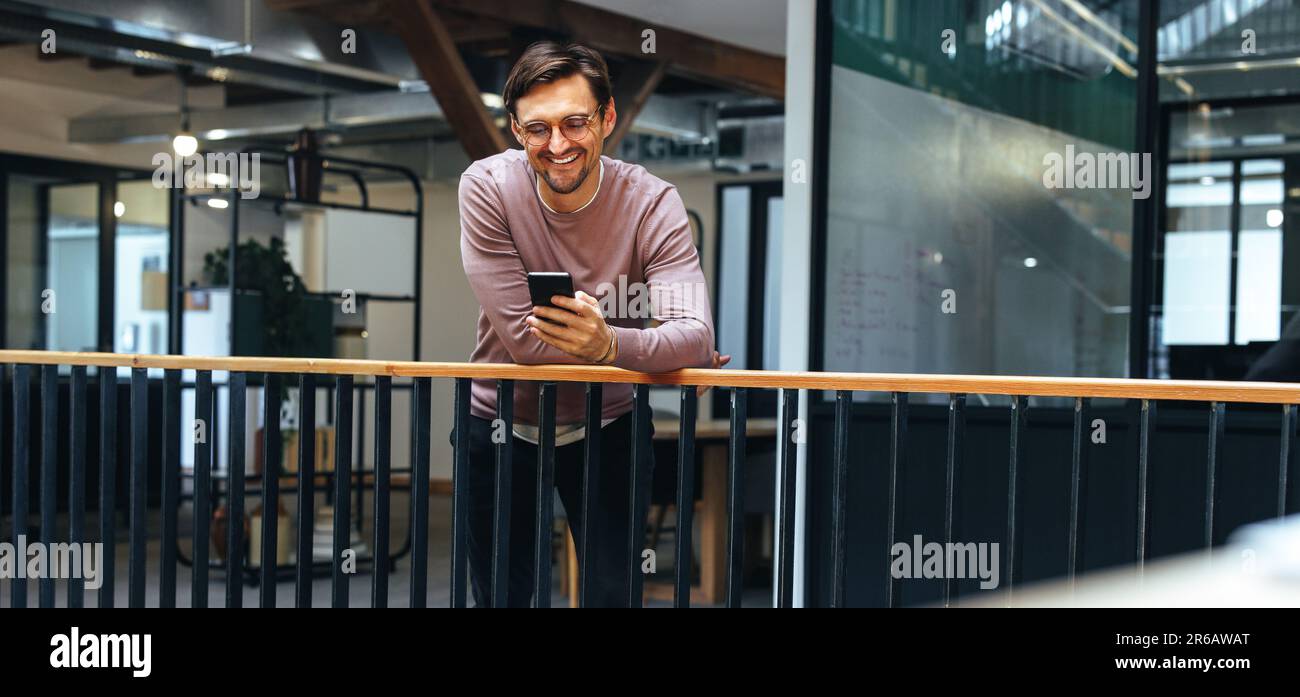 Professional man reading a text message on a smart phone in an office. Happy business man using a mobile phone while standing on an interior balcony. Stock Photo