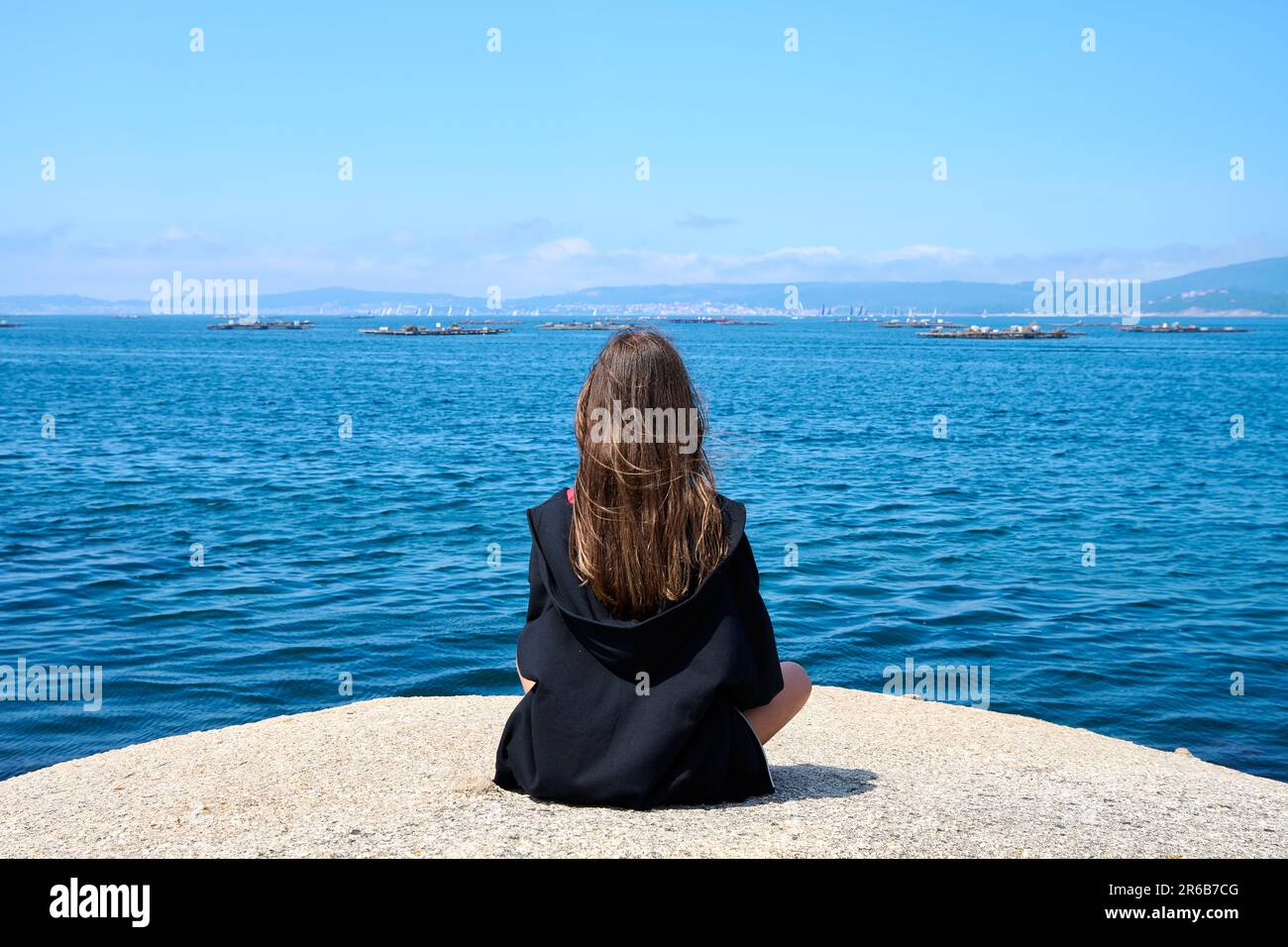 A young woman sits contentedly on the shoreline, looking out at the ...