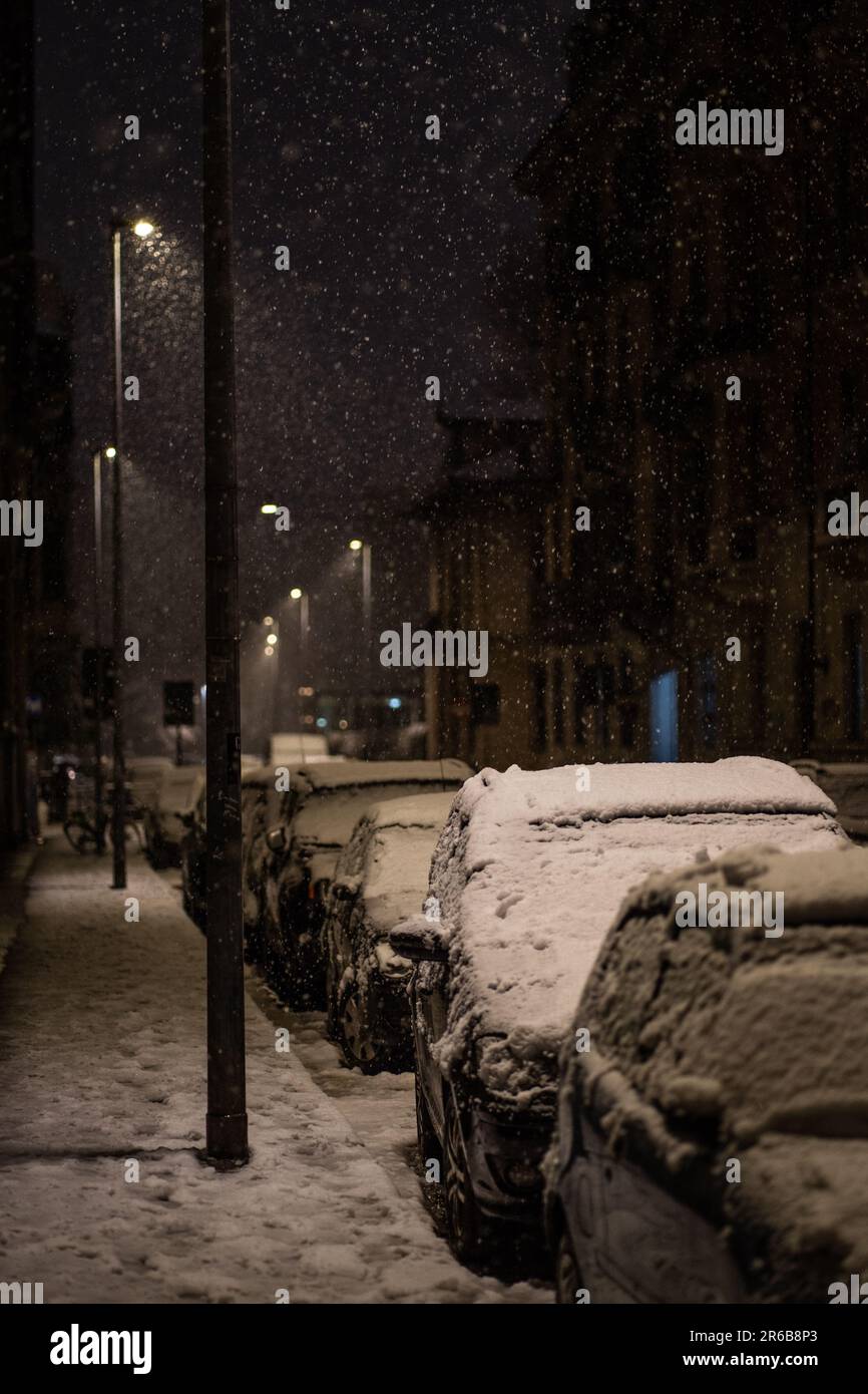 A snowy cityscape is shown with parked cars on a sidewalk and people ...