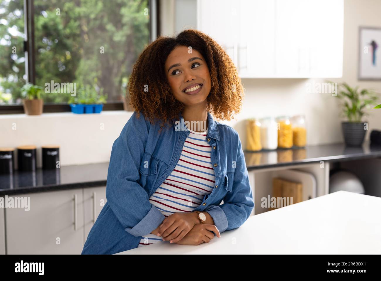 Happy biracial woman with curly hair leaning on counter smiling in kitchen Stock Photo