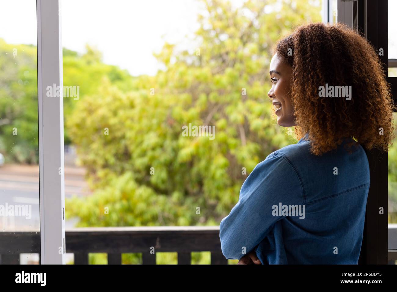 Happy biracial woman with curly hair on balcony looking at trees and smiling, with copy space Stock Photo