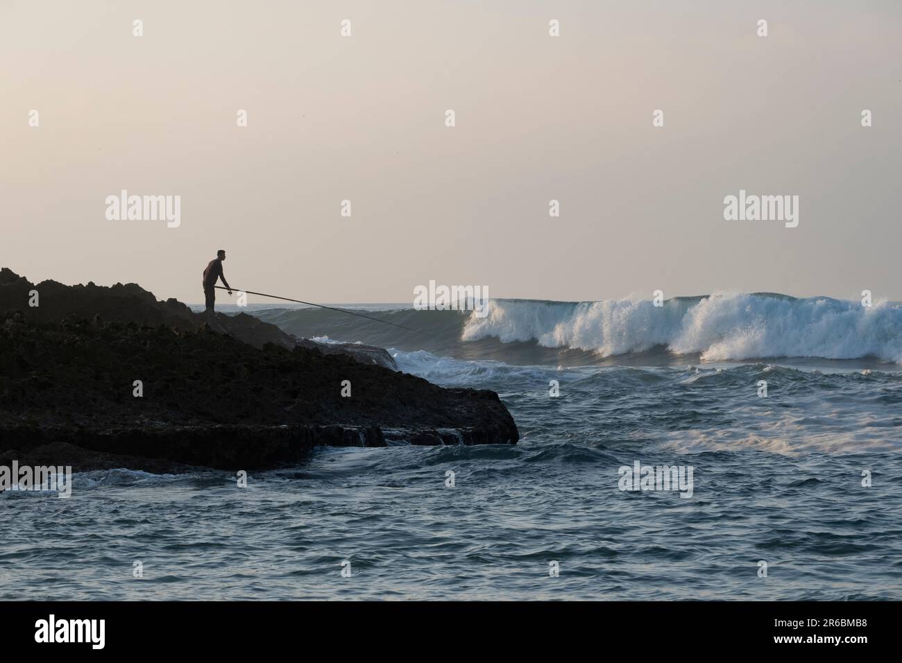 A silhouette fisherman standing on a rocky Atlantic ocean outcrop with his fishing rod pointed towards the crashing waves at Oualidia, Morocco Stock Photo