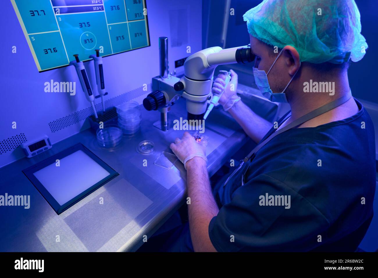 Laborant in sterile gloves and protective face mask looking at microscope Stock Photo