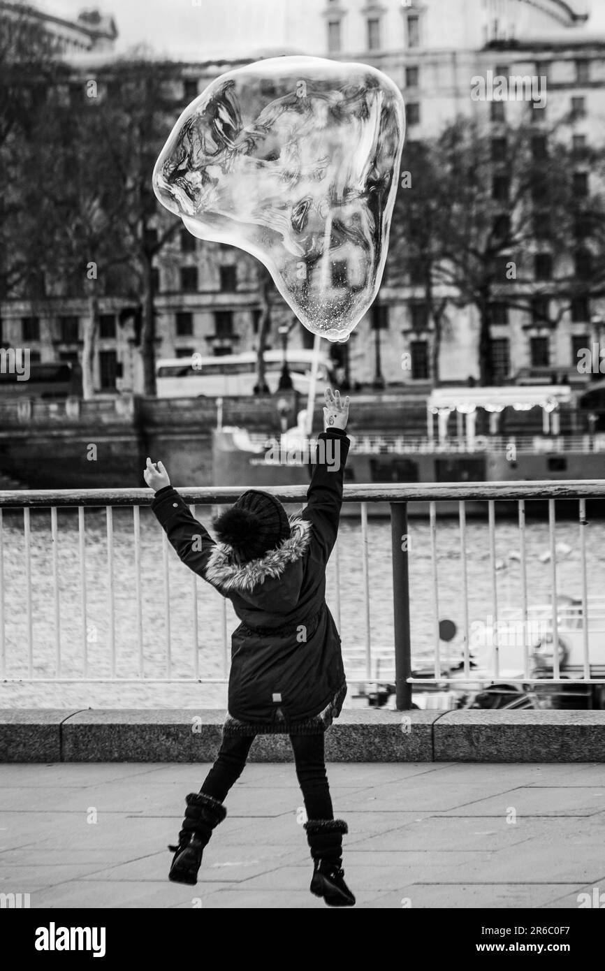 London black and white street photography: Young girl reaches to burst a large bubble floating along the Southbank, London, UK. Stock Photo