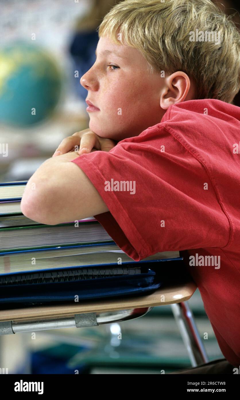 worn out boy in school not paying attention in class, leaning on a large stack of books bored student at his classroom desk Stock Photo