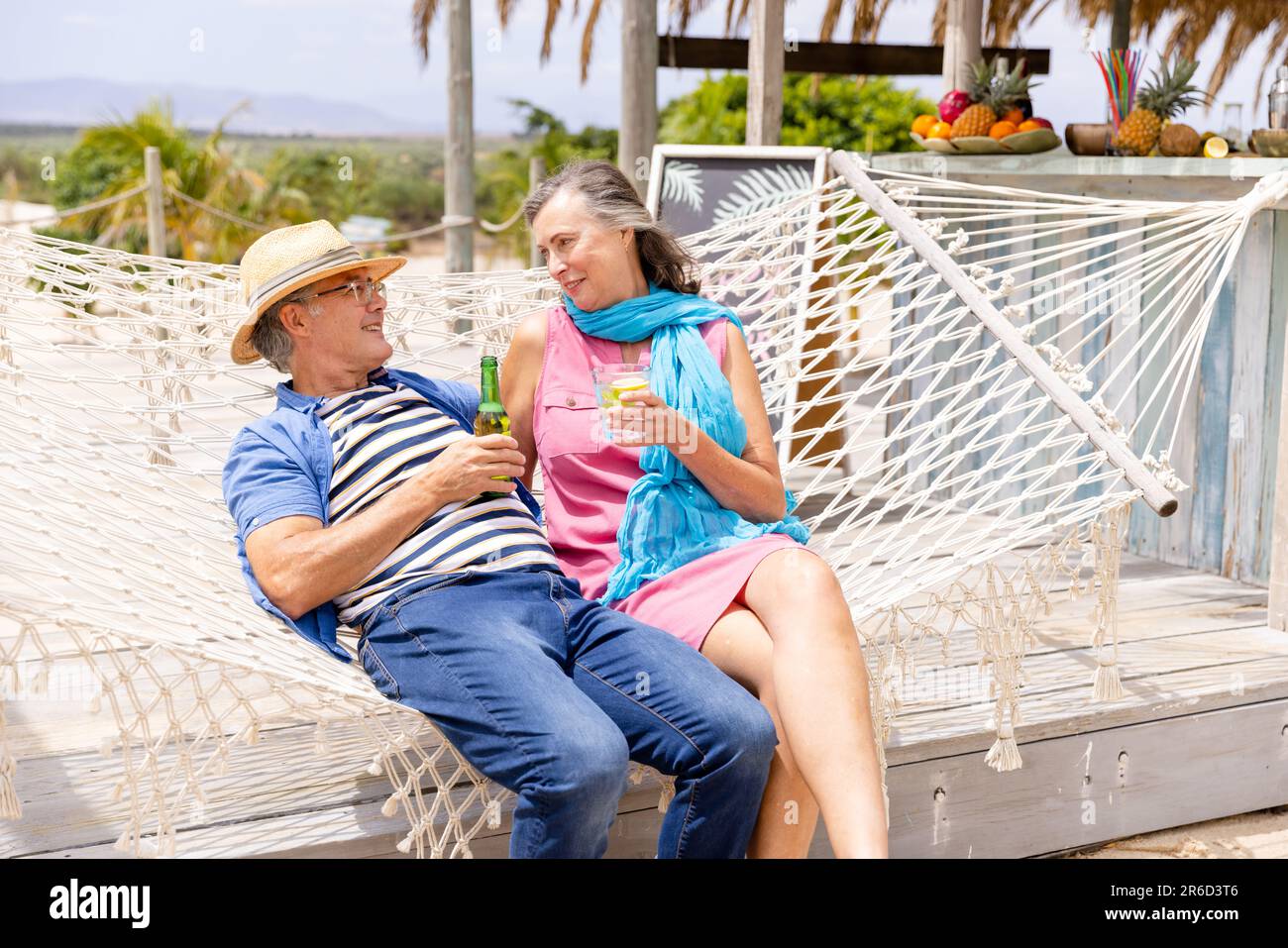 Caucasian senior couple holding drinks and talking while relaxing on hammock at beach Stock Photo