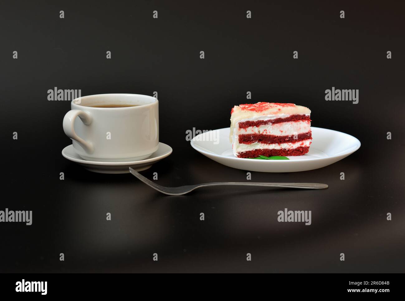 A cup of hot coffee on a saucer with a spoon and a plate with a slice of red velvet cake with mint leaves on a black background, next to a dessert for Stock Photo