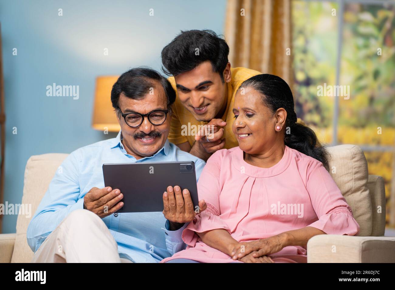 Indian senior parents with adult son watching digital tablet by talking together at home - concept of communication, parenthood and interaction. Stock Photo